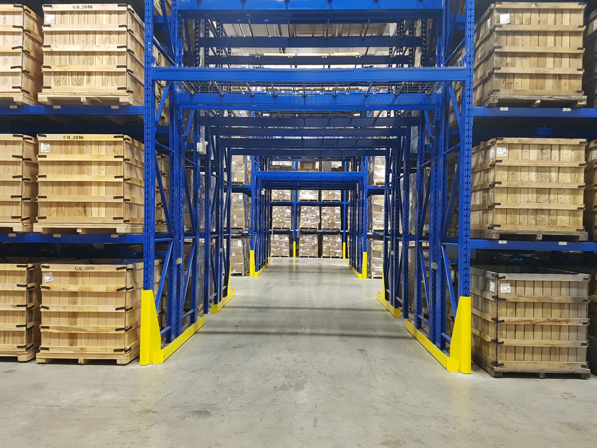 entrepot-industriel-rangement-palettes-rayonnage-metalique.png Allée centrale d’un entrepôt industriel avec rayonnage métallique bleu et palettes de bois entreposées